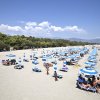 Spiaggia con ombrelloni blu e bianchi, contornata da alberi e montagne in lontananza.